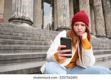 Portrait of young redhead woman with complicated face, sits on street stairs in red hat, holds smartphone and frowns thoughtful, feels uneasy. - Shutterstock ID 2257562601