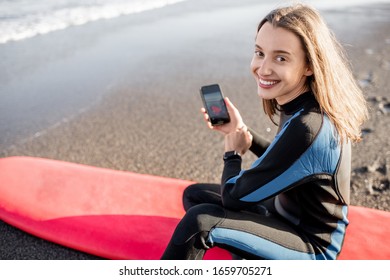 Portrait Of A Young Relaxed Surfer In Wetsuit Sitting With Surfboard And Smart Phone On The Beach. Water Sport And Leisure Activity Concept