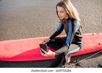 Portrait Of A Young Relaxed Surfer In Wetsuit Sitting With Surfboard And Smart Phone On The Beach. Water Sport And Leisure Activity Concept