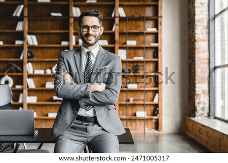 Portrait of young rich businessman in formal suit with arms crossed looking at the camera in modern office. Wealthy foreman manager working, making money at workplace