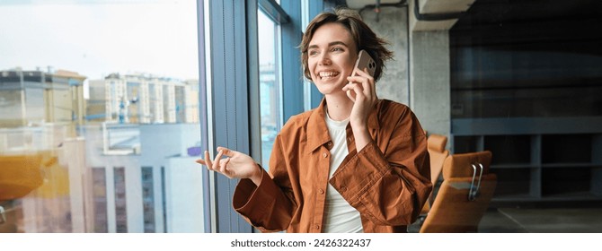 Portrait of young saleswoman, office worker calls client, businesswoman talking on mobile phone and standing near window at workplace. - Powered by Shutterstock