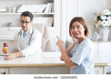 Portrait Of Young Scared And Irritated Female Patient And Doctor Cardiologist Working In Office Of Modern Clinic	
