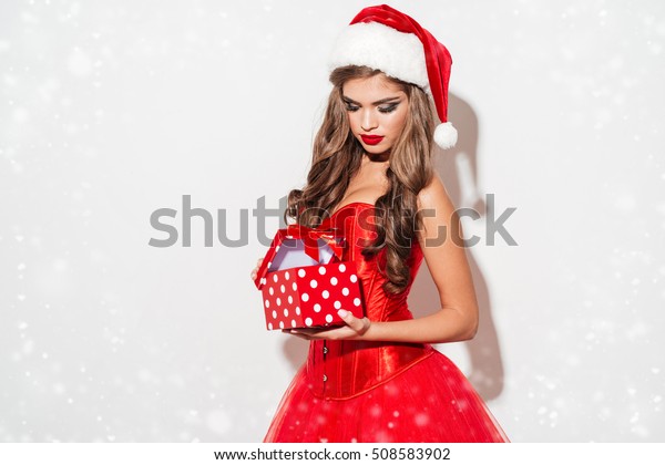 Portrait of a young seductive brunette woman in red santa claus outfit opening small gift box isolated on the white background