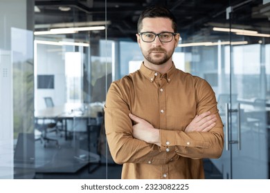 Portrait of a young self-confident businessman, executive director, founder standing in an office building. He crosses his arms on his chest and looks seriously into the camera. - Shutterstock ID 2323082225