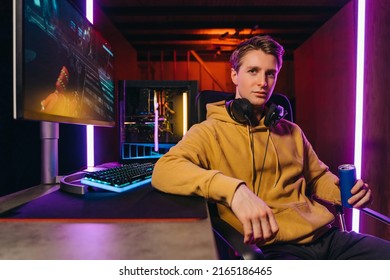 Portrait of young serious handsome pro gamer sitting at desk with gaming equipment, holding energy drink, using powerful PC for playing online video shooter games. Neon Colored Room, e-Sport concept 