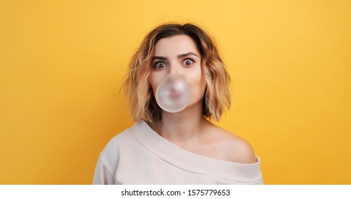 Portrait Young Sexy Girl Diligently Inflates Bubble Of Chewing Gum And Rounds Her Eyes From The Effort On An Isolated Yellow Background. Pleasure. The Emotions Of People. Copy Space. Lifestyle