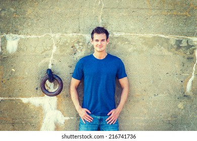 Portrait Of Young Sexy Man.  Wearing A Blue T Shirt, Jeans, A Young Handsome Guy Standing Against An Old Concrete Wall With A Rusty Metal Ring, Looking At You. Concept Of Timing, History And Life.