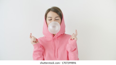 Portrait Of Young Sexy Playful Girl Dressed In Pink Hoodie Standing Over White Background Blowing Bubblegum Candy Bubbles And Chewing Gum. Woman Looking At Camera