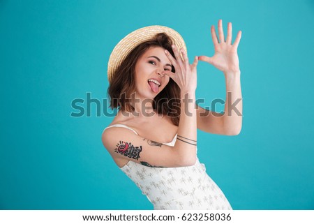Portrait of a young silly beach girl wearing summer clothes and teasing with hands at her nose isolated over blue background