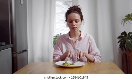 Portrait Of Young Skinny Woman Sitting At Table And Eating Piece Of Cucumber And Salad With For And Knife. Female On Severe Diet Starving And Having Vegetables For Dinner