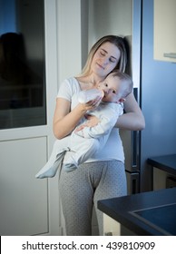 Portrait Of Young Sleepy Mother Feeding Baby Son From Bottle On Kitchen At Night