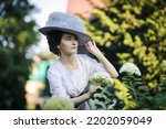 Portrait of a young slender woman in a 1910s costume. A lady in a hat in the fashion of the early 20th century walks in a garden and looks at hydrangea flowers