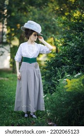 Portrait of a young slender woman in a 1910s edvardian costume. A lady in a hat and glasses in the fashion of the early 20th century walks in a garden