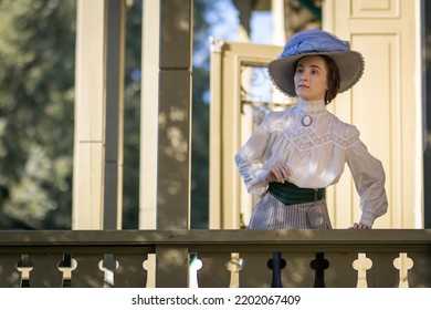 Portrait of a young slender woman in a 1910s costume. A woman in an Edwardian hat, blouse and skirt on the veranda of the estate.