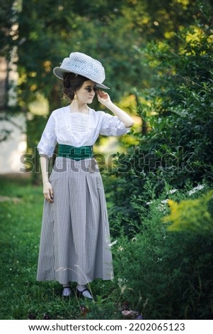 Portrait of a young slender woman in a 1910s edvardian costume. A lady in a hat and glasses in the fashion of the early 20th century walks in a garden