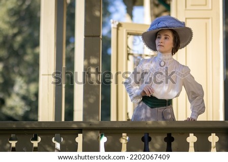 Portrait of a young slender woman in a 1910s costume. A woman in an Edwardian hat, blouse and skirt on the veranda of the estate.