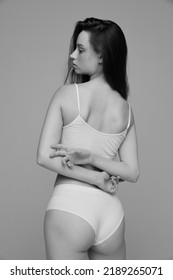 Portrait Of Young Slim Girl With Dark Hair Posing In White Underwear. Black And White Photography Aesthetics. Healthy Back. Concept Of Natural Beauty, Body And Skin Care, Femininity, Self-acceptance