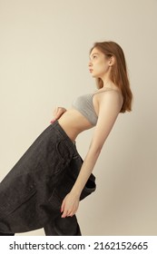 Portrait Of Young Slim Girl In Top And Giant Black Jeans Posing Isolated Over Grey Studio Background. Weight-loss. Anorexia. Concept Of Beauty, Health, Body And Skin Care, Special Diet,, Fitness, Ad