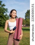 Portrait of young slim and healthy korean girl doing workout in park, standing with water bottle and rubber mat for execises on green lawn, smiling happily.