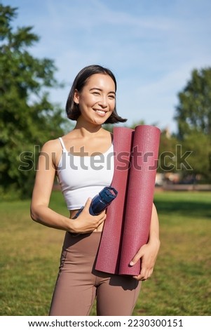 Portrait of young slim and healthy korean girl doing workout in park, standing with water bottle and rubber mat for execises on green lawn, smiling happily.