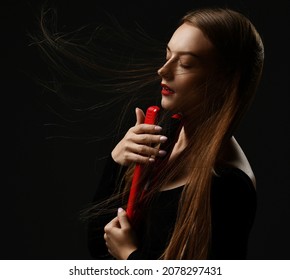 Portrait of young slim sensual woman with silky fluttering in the wind hair and eyes closed holding red hair straightener at face over dark background. Haircare, beauty, wellness concept