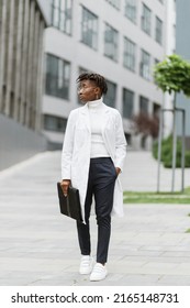 Portrait Of Young Smart Focused African Female Medical Intern Doctor With Folder Clipboard, Standing Outside Modern Hospital Building. Medicine, Education And Healthcare Concept.