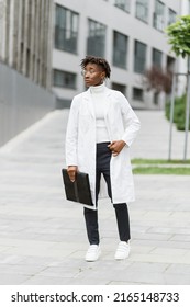 Portrait Of Young Smart Focused African Female Medical Intern Doctor With Folder Clipboard, Standing Outside Modern Hospital Building. Medicine, Education And Healthcare Concept.