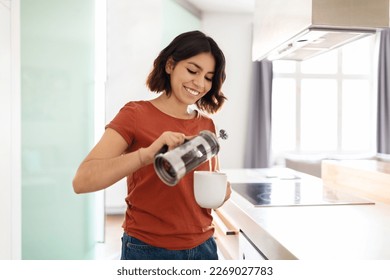 Portrait Of Young Smiling Arab Woman Making Morning Coffee At Home, Happy Millennial Middle Eastern Female Standing In Kitchen Interior, Holding French Press And Pouring Caffeine Drink To Cup