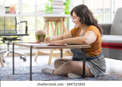 Portrait Of Young Smiling Asian Fat Female Sitting On A Floor Studying And Writing On A Book On A Table In A House With Delightful. Study From Home Concept. Focus On A Chubby Woman Face.