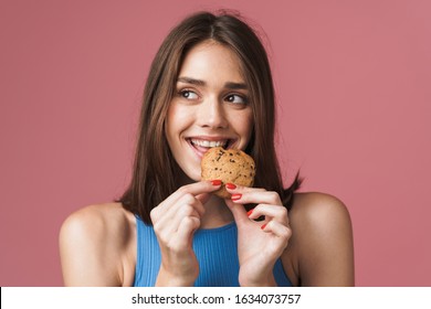 Portrait Of A Young Smiling Attractive Brunette Woman Standing Isolated Over Pink Background, Eating A Chocolate Chip Cookie