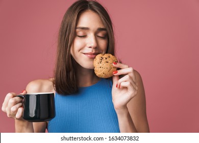 Portrait Of A Young Smiling Attractive Brunette Woman Standing Isolated Over Pink Background, Holding A Cup Of Coffee And Eating A Chocolate Chip Cookie