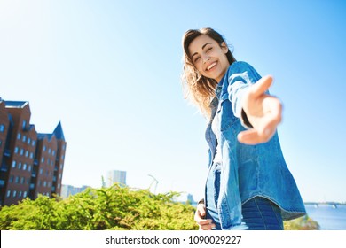portrait of a young smiling attractive woman in jeans clothes at sunny day on the blue sky background. happy woman gives a hand to someone like follow me . first person view.