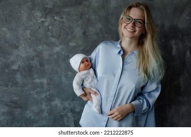 Portrait Of Young Smiling Beautiful Woman Wearing Blue Shirt, Holding Baby Doll In White Clothes On Grey Marble Background. Motherhood, Childbirth, Breastfeeding, Breast Model, Fake, Education. 