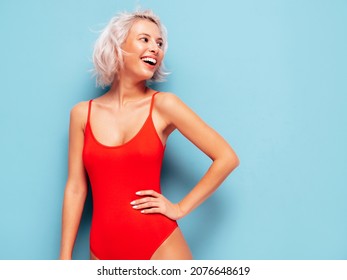 Portrait of young smiling blond model in summer swimwear red bathing suit. Sexy carefree woman having fun and going crazy. Female posing near blue wall in studio. Cheerful and happy.