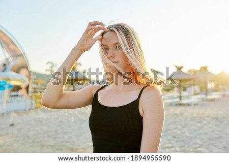 Portrait of young smiling blonde teenager in black top on the beach, sunset sandy seashore background
