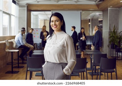 Portrait of young smiling brunette professional business coach woman in office looking at camera. Students, group of people on background. Independent, confident woman stands with hands in pockets. - Shutterstock ID 2266968405