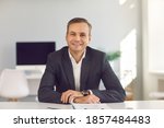 Portrait of young smiling businessman in suit sitting and looking at camera during online videocall in office over modern company interior at background. Working in office, business, online concept
