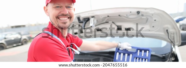 Portrait of young smiling car repairman with set of\
tools on hood of car