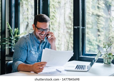 Portrait of young smiling cheerful entrepreneur in casual office making phone call while working with charts and graphs looking at paper documents - Shutterstock ID 1477845698