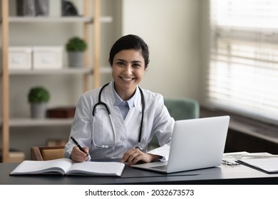 Portrait Of Young Smiling Confident Indian Ethnicity Female Doctor General Practitioner Therapist In Medical Uniform Working On Computer, Making Records In Modern Clinic Office Room Sitting At Table.