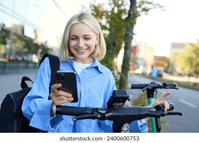 Portrait of young smiling female model, renting an electric scooter, using mobile phone to scan QR code with smartphone app, riding home from university, standing outdoors on street. - Powered by Shutterstock