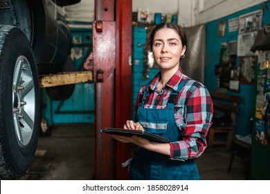 Portrait Of A Young Smiling Female Worker In Uniform Posing With A Tablet In Her Hands. In The Background Is An Auto Repair Shop