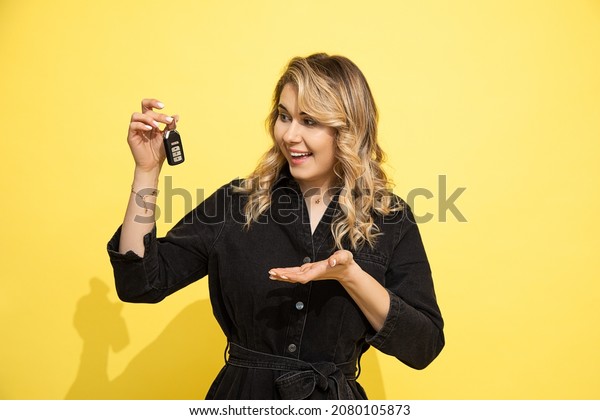 Portrait of\
Young smiling happy satisfied excited successful blonde woman in\
black denim dress holding in hand and pointing with finger car keys\
on yellow background, studio photo shot.\
