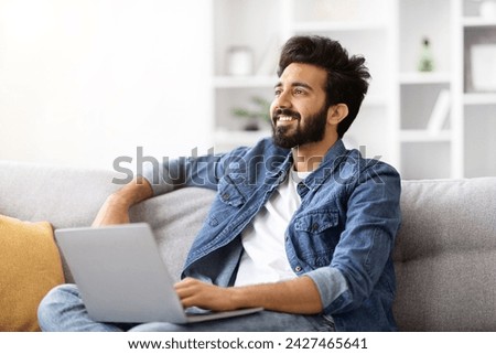 Portrait Of Young Smiling Indian Man With Laptop Relaxing On Couch At Home, Dreamy Millennial Eastern Guy Sitting With Computer On Sofa In Cozy Living Room And Looking Away, Copy Space