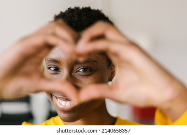 Portrait of young smiling lovely multiracial woman showing shape heart with hands and looking through it with toothy smile while posing at her home - Love and happy people concept