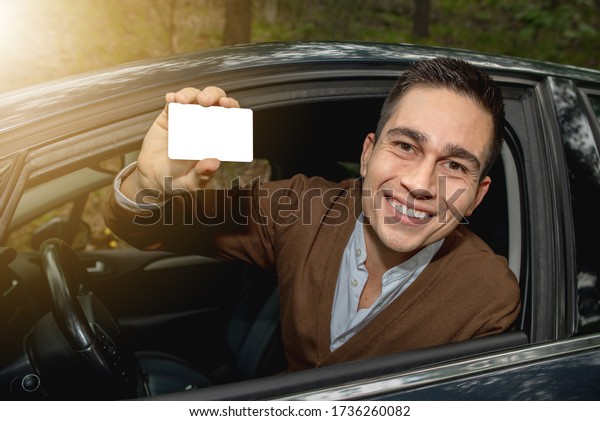 Portrait of young smiling man inside his new\
car posted outside the window showing the driver license or other\
document, free space for text in the\
card.