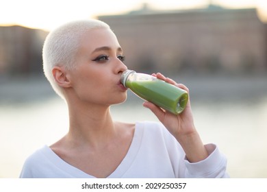 Portrait Of Young Smiling Millenial European Short Haired Woman Drinking Green Smoothie In Bottle. Beautiful Happy Blonde Girl Outdoor.