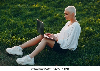 Portrait Of Young Smiling Millenial European Short Haired Woman Using Laptop At Green Grass Meadow In Park. Beautiful Happy Blonde Girl Outdoor. Summer Fashion Female Clothing.