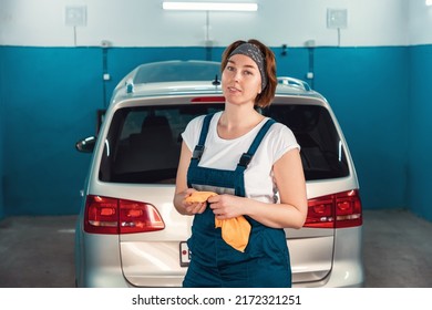 Portrait Of A Young Smiling Plus-sized Woman In Overall, Wiping Her Hands With A Rag. At The Background Of A Car In An Auto Repair Shop. The Concept Of Women's Equality And Work In Garage.