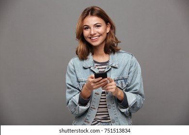 Portrait of a young smiling teenage girl dressed in denim jacket holding mobile phone and looking at camera isolated over gray background